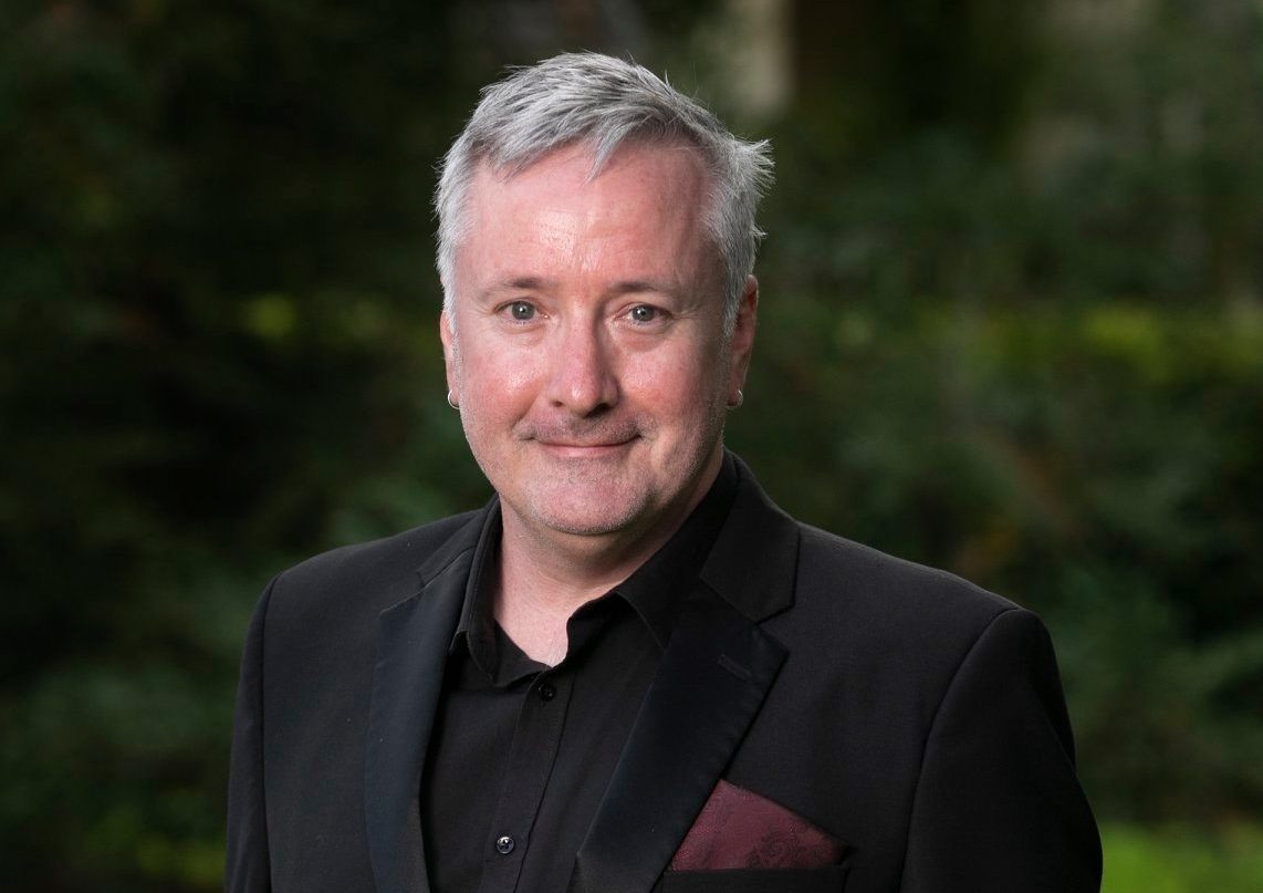 A grey-hair Anglo-Australian man wearing a black suit with a dark red pocket square smiles at the camera. Behind him is a green lawn and trees.