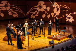 Perfumes of the East. SXS performers stand on stage with their instruments. A soloist stands in a green dress. Large flowers adorn the side and back of the stage.