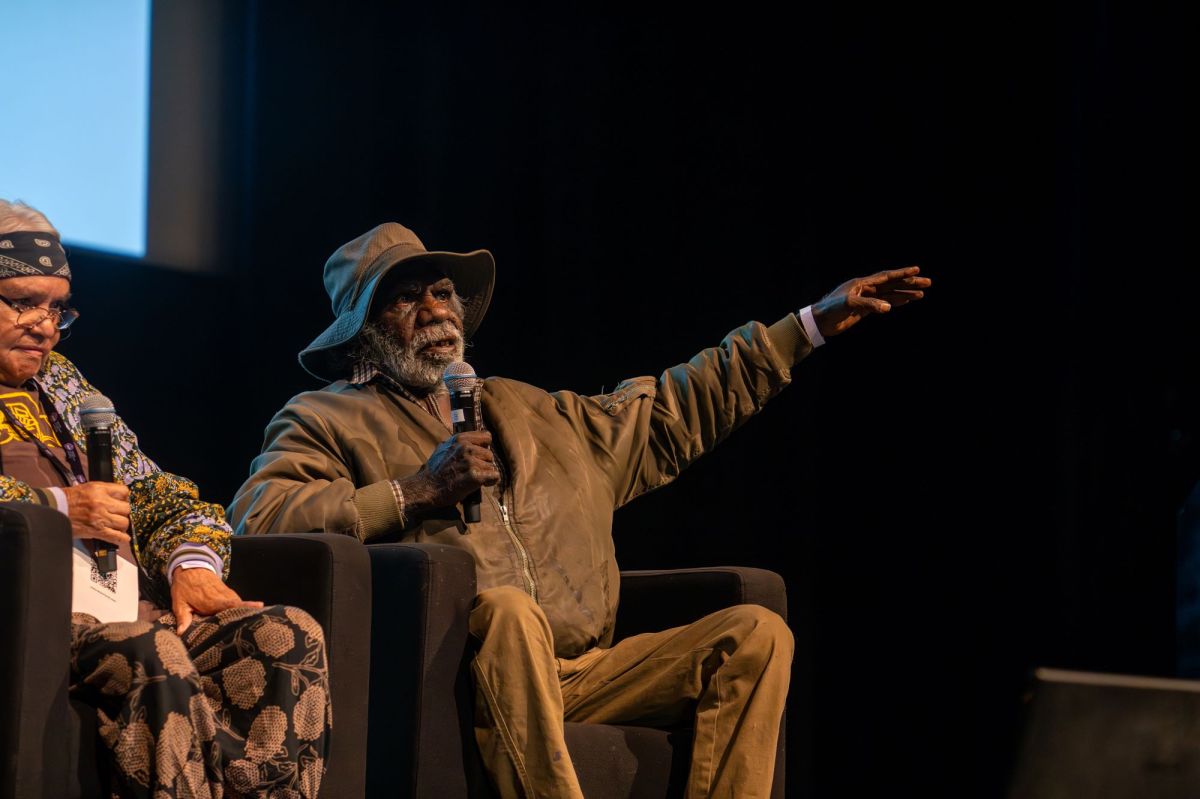 Selma Coultard and Mervyn Rubuntja at the Desert Mob Symposium 2023. Photo: Rhett Hammerton. A dark-skinned Aboriginal man with a short grey beard gestures with his left hand while holding a microphone in his right hand, into which he is speaking. He wears a brown hat, brown jacket and tan-coloured slacks. A brown-skinned Aboriginal woman wearing glasses, with her hair hair held back by a headscarf, sits to his right, but she is not the main focus of the photograph. The two sit beneath a screen, suggesting they are speaking on stage together.