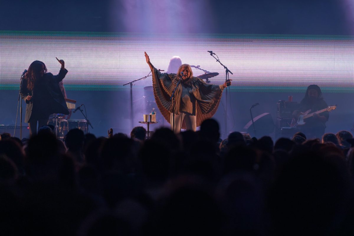 A throng of punters watch on as three musicians perform on stage. Two of them are indistinct on the dark stage but Asha Puythi is well lit. With her arms outstretched, her colourful poncho resembles butterfly wings.