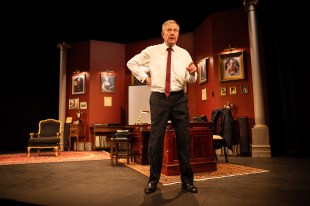 A man in a white shirt, black pants and tie is standing in front of an office set. He's got on hand on his hip and another pointing at the viewer.