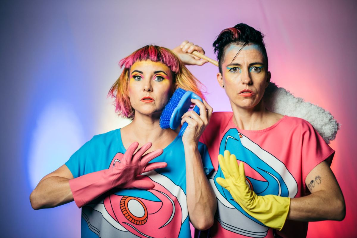 Two female-presenting individuals pose for the camera. They both have a colourful 1980s-inspired look, as does the background of the photo. One wears a pink rubber glove and holds a blue scrubbing brush up to her eat like a telephone. The other wears a single yellow rubber glove and wields a feather duster like a sword. They are The Ironing Maidens, who are performing at Riverfest X.