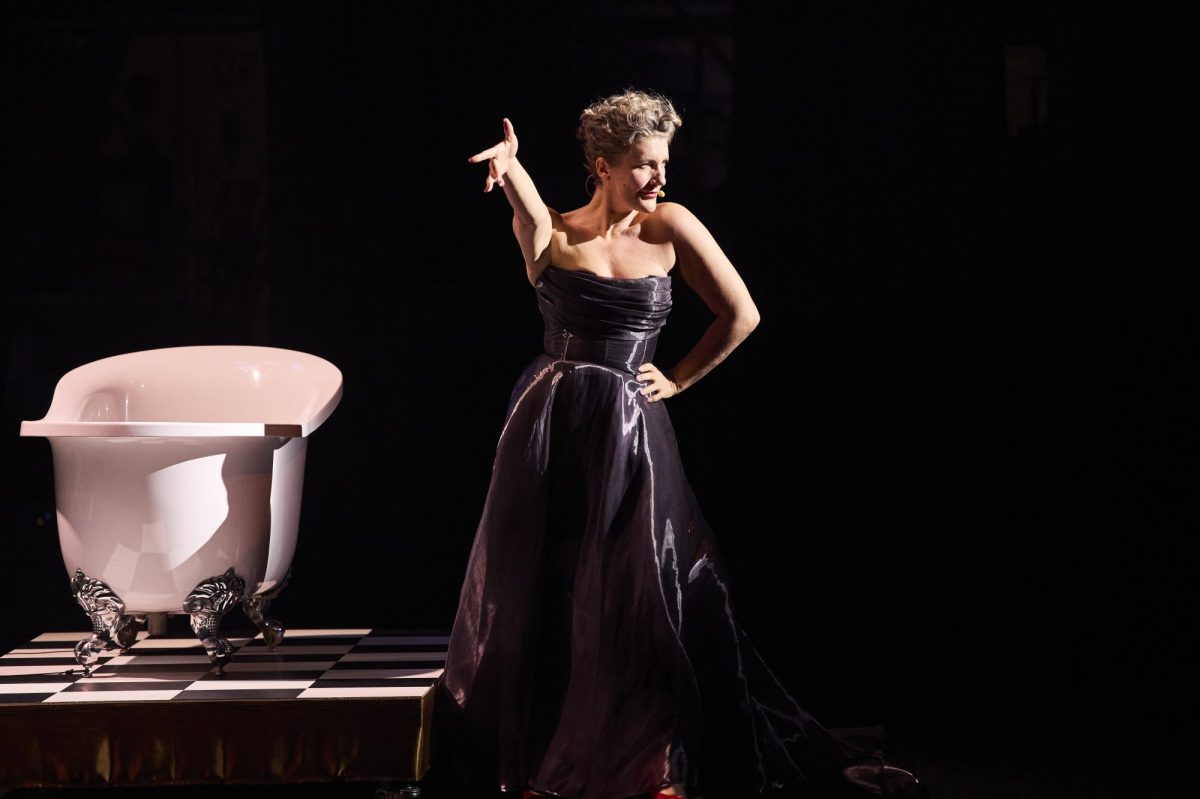 A blonde woman in a strapless evening gown is standing next to a bathtub, which is on a raised platform.