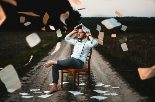 Man sitting on chair outside with paper pages raining down on him. Arts news.