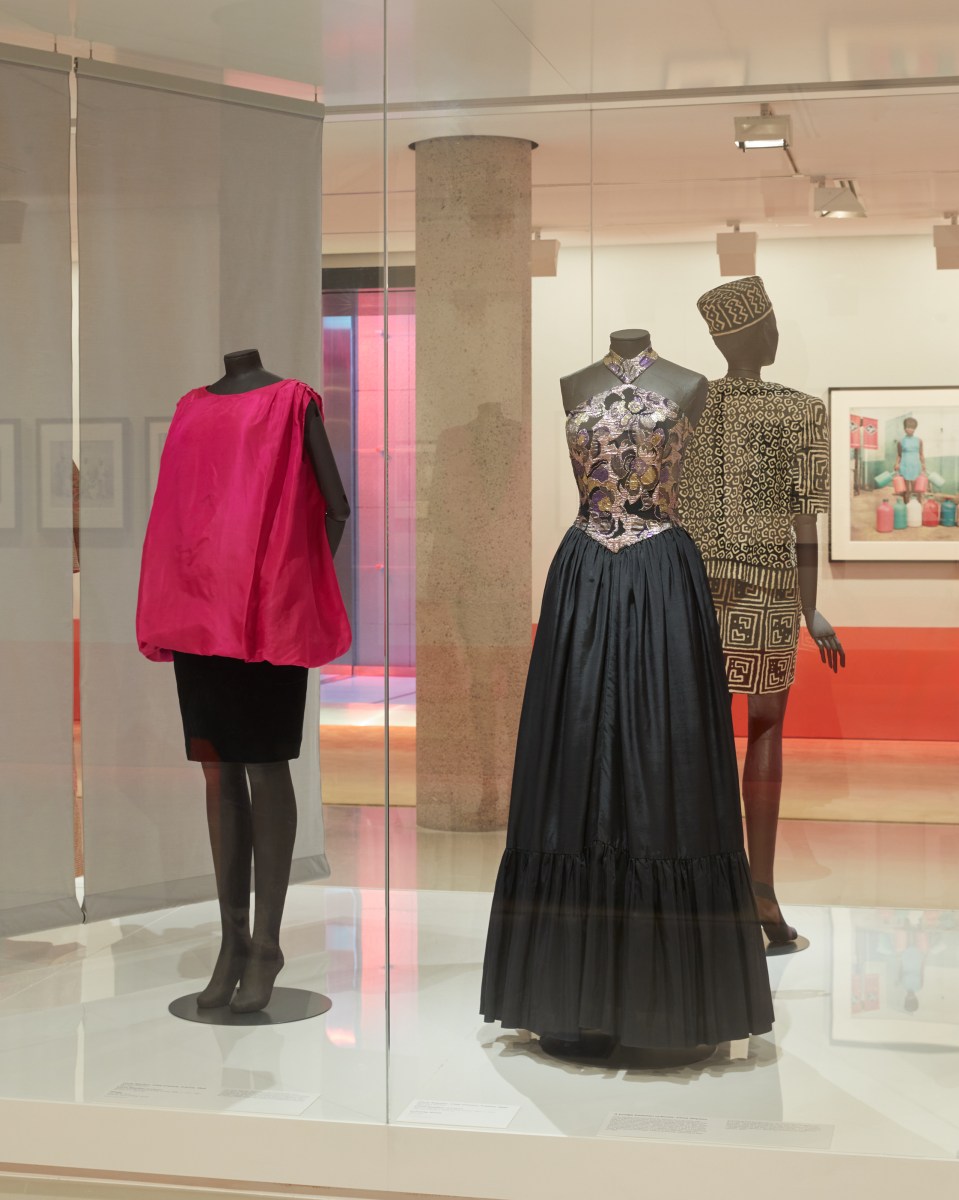 Installation view of ‘Africa Fashion’ on display from 31 May to 6 October 2024 at NGV International, Melbourne. Photo: Lillie Thompson. Three garments in a glass display, one featuring a balloon sleeveless pink top and black skirt, another with a purple embroidered dress and a third (back facing) gold and black suit set. 