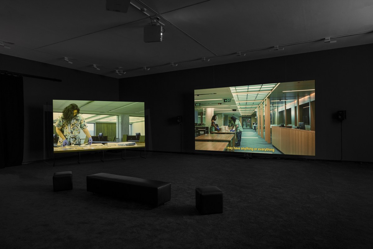 Andy Butler, 'Living truthfully in invented circumstances', 2024, installation view at ACCA. Photo: Andrew Curtis. Two video projections, one smaller and one larger in a dark gallery space with three black benches placed in front. Both video imagery feature people inside an archival space, sorting through material.