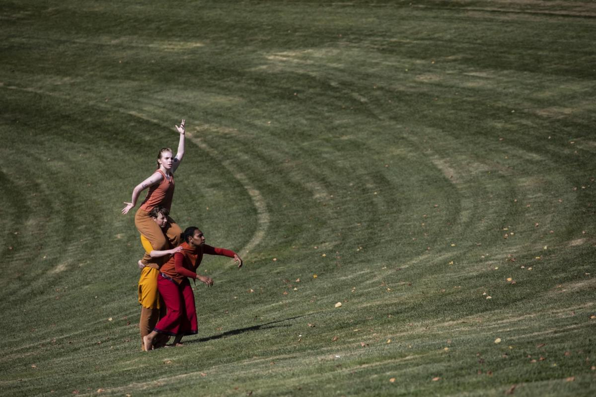 Three women are out in a field. One is carrying another one on her shoulders. They are mid-dance. They are wearing flame-coloured clothing.