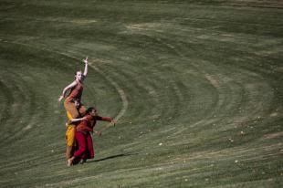 Three women are out in a field. One is carrying another one on her shoulders. They are mid-dance. They are wearing flame-coloured clothing.