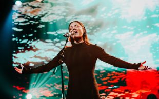 2024 Australian Women in Music Awards finalists announced. Image: Vanessa Amorosi performing at AWMA 2023. A women with long brown hair singing on stage with her eyes closed and arms outstretched. The background appears to be a projection of light shining across a body of water.