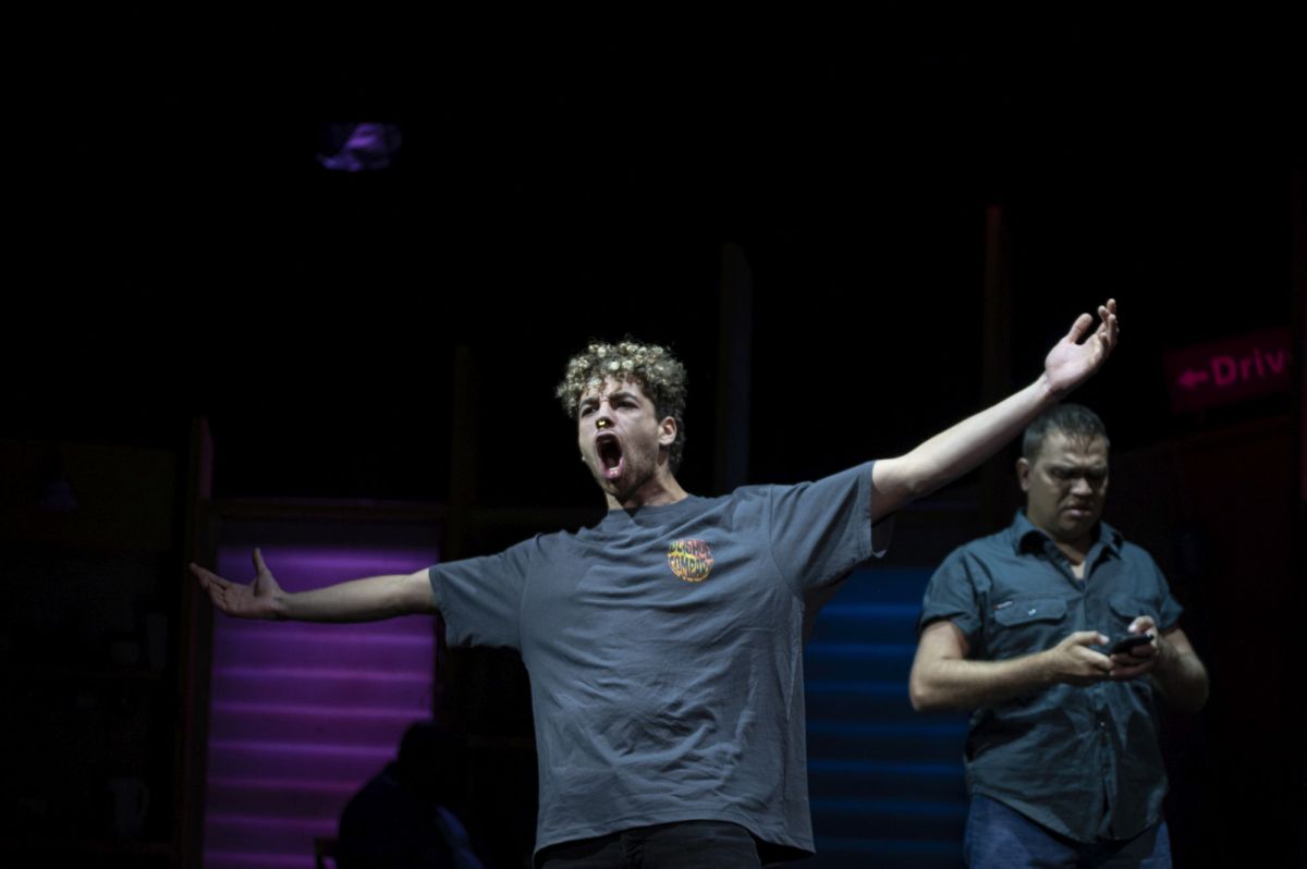 Two young men wearing blue short-sleeve tops. One is in the centre of the frame with his arms wide apart and his mouth open. The other is standing on the right side looking down at his phone.