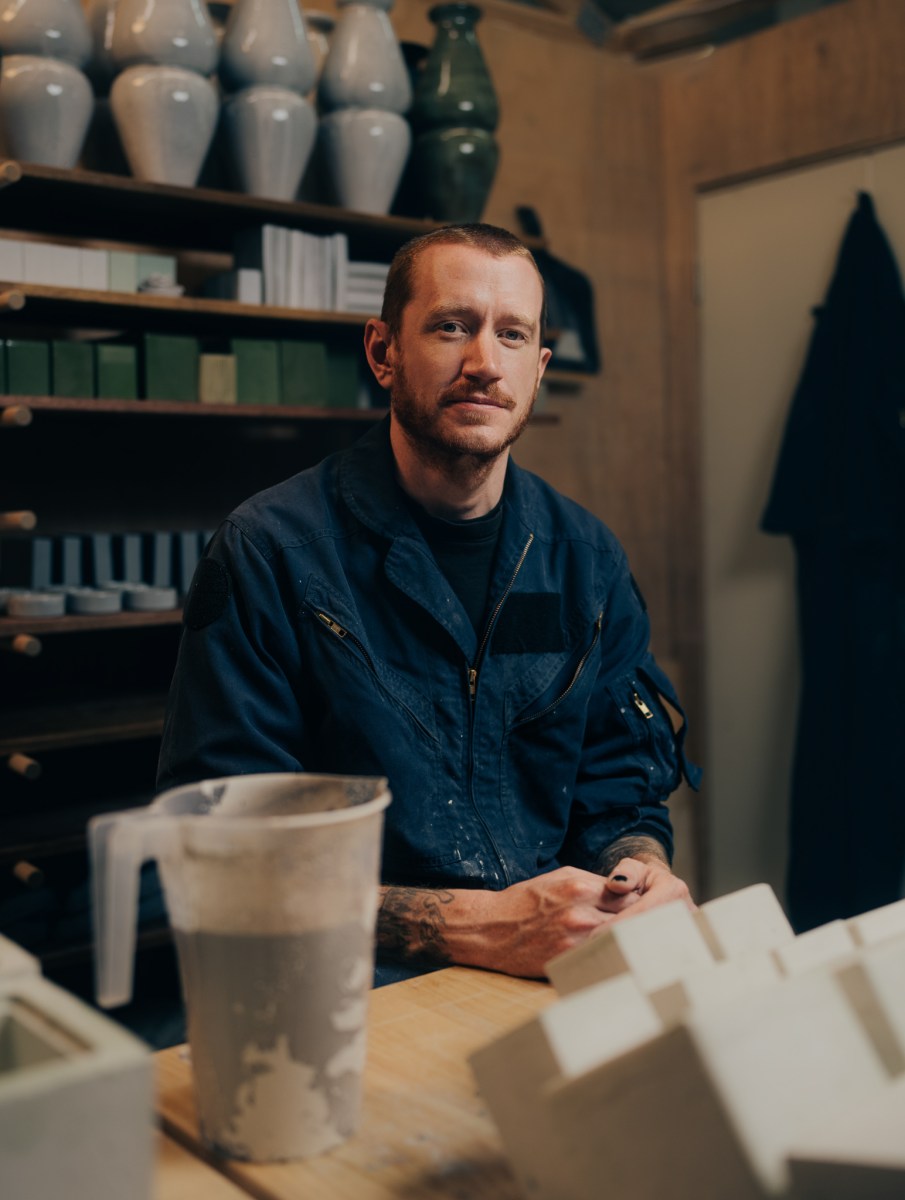 Stanthorpe. A young Caucasian man with short hair and a moustache sits in a blue shirt next to blocks of artist's clay and a jug. There are finished pieces of ceramics behind him on shelves.