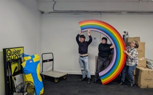 Midsumma on the move, destination: unknown. A photo of three people moving a rainbow prop inside an office space with white walls and grey carpet. There is a trolley nearby and other storage times such as a signage with the word ‘Queer’.