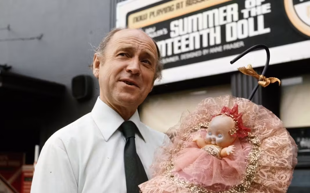 Tay Lawler, a balding, older Anglo-Australian man wearing a white shirt and tie, stands in front of a theatre advertising the 1977 revival of 'Summer of the Seventeenth Doll'. He is holding a pink Kewpie Doll, like those featured in the play.