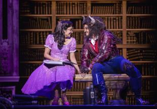 A woman with brown hair and a pink dress and an open book on her lap is sitting next to a man dressed up as a beast, with horns and facial hair. There is a backdrop of a library.