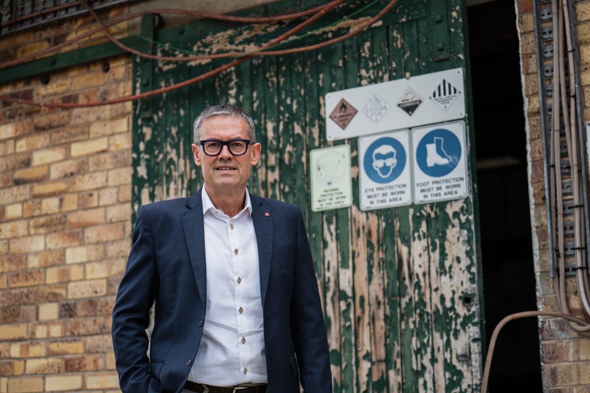 A middle aged Caucasian man with grey hair and black framed glasses, white shirt and suit jacket stands in front of a paint peeling doors with industrial signs on it.
