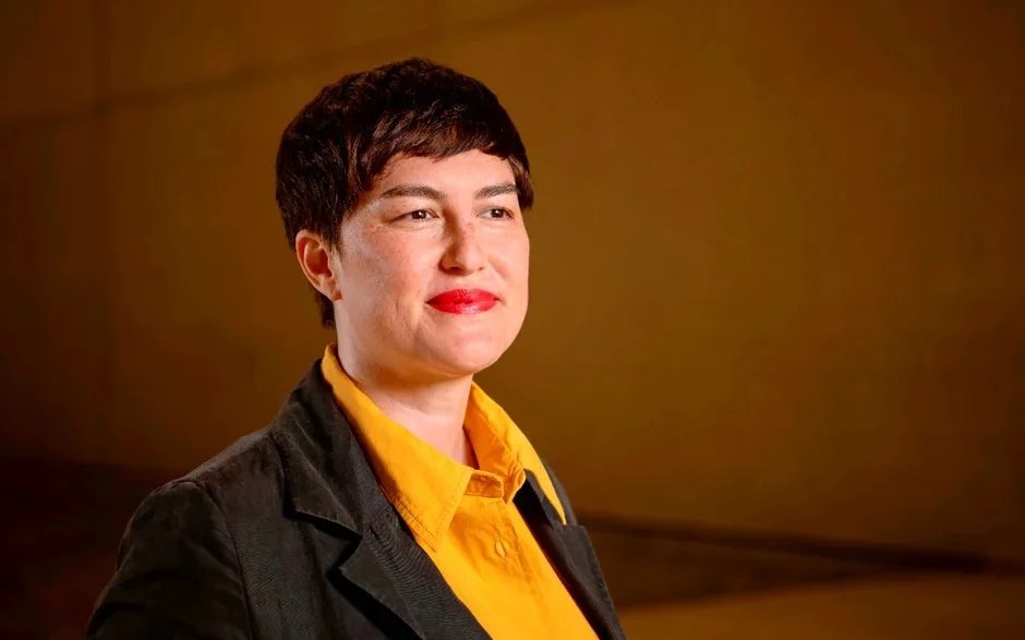 2026 Adelaide Biennial curator Ellie Buttrose. A middle-aged woman with pale skin and short cropped brown hair, wearing red lipstick, an orange shirt and black blazer. She is looking off to the side of the camera and smiling.