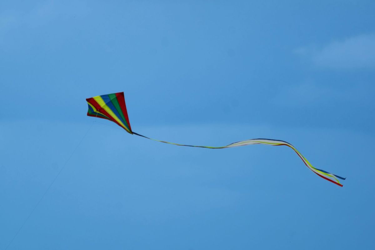 A colourfully-striped triangular kite with a long, bright tail flies against a clear blue sky.