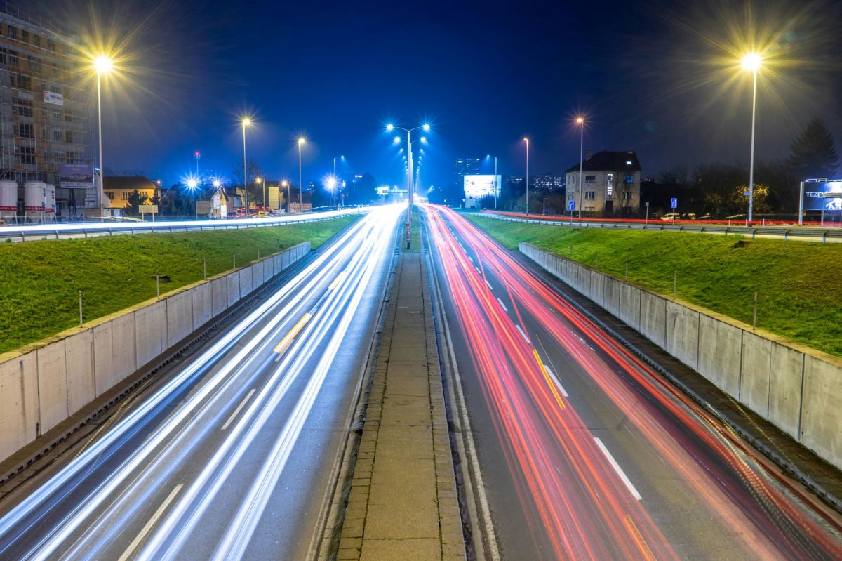 An urban roadway at night, with the blurred headlights and taillights of cars painting stripes of colour as they move towards and away from the camera.