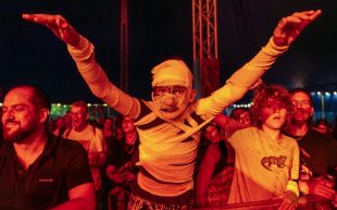 Festival-goers at Byron Bay Bluesfest. A crowd of people gathering below a stage facing the camera. A figure in the centre of the photo is wrapped up like a mummy with their arms outstretched.