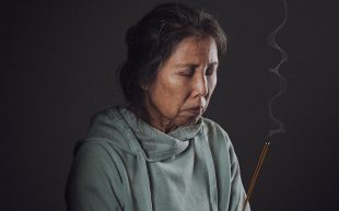 Anna Yen plays Mrs Chen in ‘AI May’, an elderly migrant mother grappling with grief in an increasingly lonely society. An elderly Asian woman wearing a sage coloured hoodie is holding a burning incense in her hands, she appears to be praying with her eyes closed.