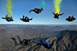 A dynamic photograph of seven sky divers, some holding flares, as they begin to link up while in freefall above a rocky landscape.