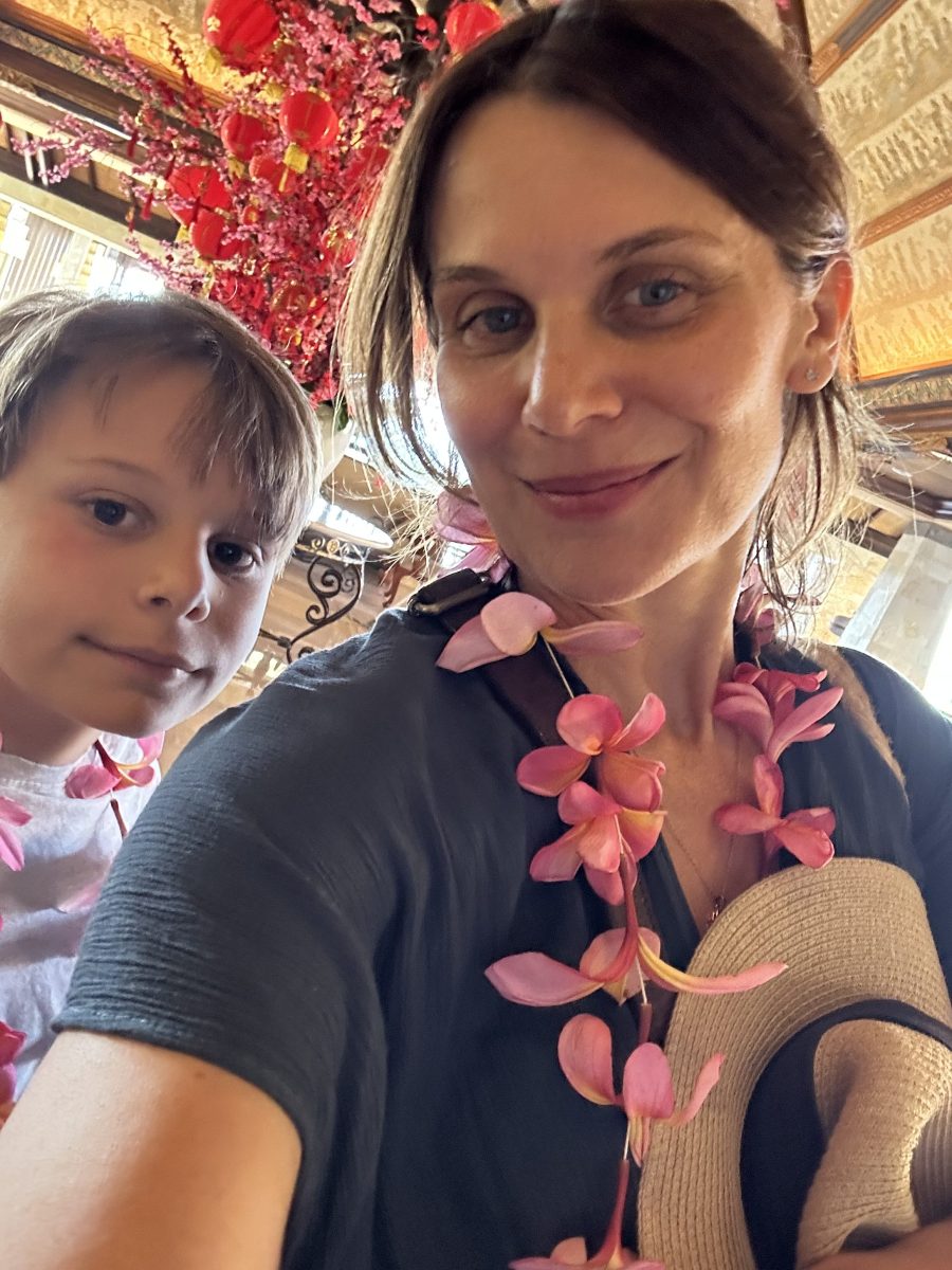 Diana Glenn and son. Image is a middle aged fair-skinned woman with brown tied back hair, a grey T shirt and a pink flower garland around her neck, holding a straw hat in front of her. Peering over her shoulder is her young son.