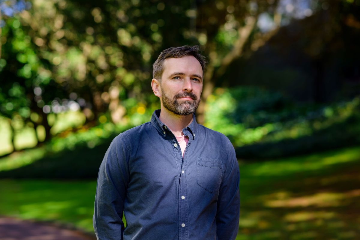 A bearded man in a blue open necked shirt stands in a park with his hands behind his back. Joel Baxendale.