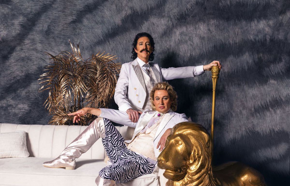 A carefully staged and rather camp photograph of two men dressed in white and lounging on a white couch. A statue of a golden lion sits beside one of the men, who are the stars of Sydney Festival's 'Siegfried & Roy: The Unauthorised Opera'.