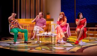 Two men and two wome are seated. All are drinking. All are wearing colourful clothes. The design colours are bright graphic prints.