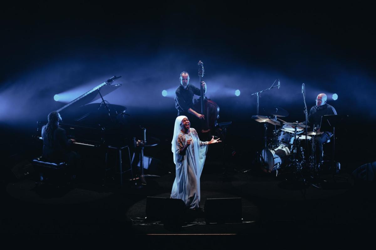 Jazzmeia Horn in white is surrounded by a band: a pianist, a double bass player and a drummer. The stage is lit in dark blue.