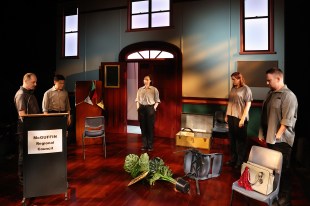 The cast of 'McGuffin Park', two women and three men, in grey clothing are in a building with a fallen fake plant. On the left is a sign attached to a lectern that says 'McGuffin regional council.'