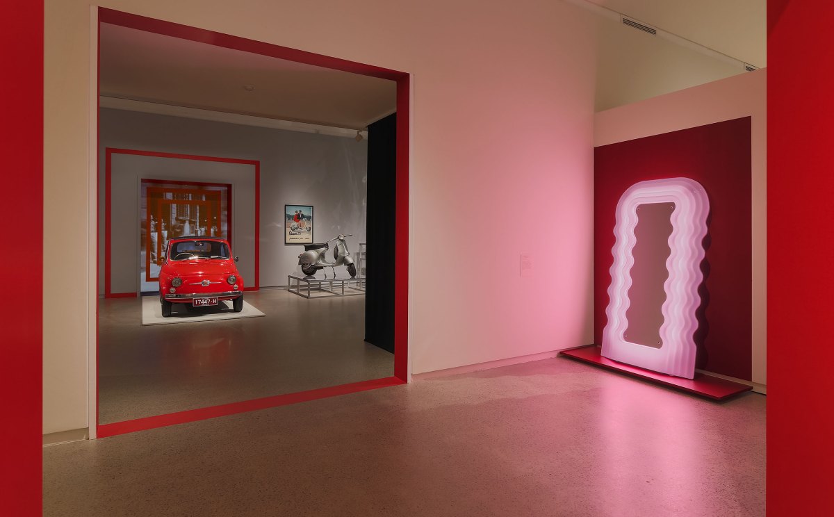 'Molto Bello: Icons of Modern Italian Design', installation view at Heide Museum of Modern Art. A gallery with white walls and red lining, featuring a red Fiat 500 car, a silver vespa, and a pink mirror with wiggly lines to the right hand side.