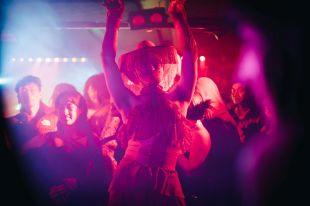 Asia TOPA's newly announced Nightlife program is a dynamic blend of performance art and club culture. A person wearing a fringed cowboy hat, facemask and fringed and laced-up dress poses dramatically, arms raised, before a culturally diverse crowd of young adults.