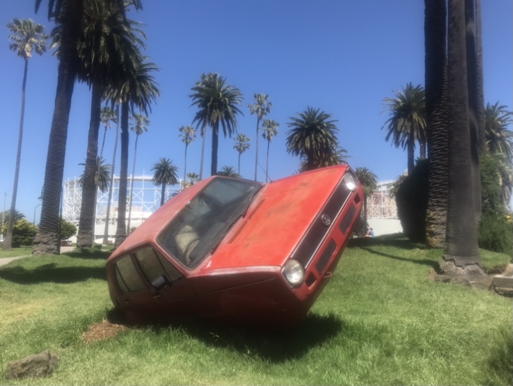 A red car installation that is perched on one side on grass.