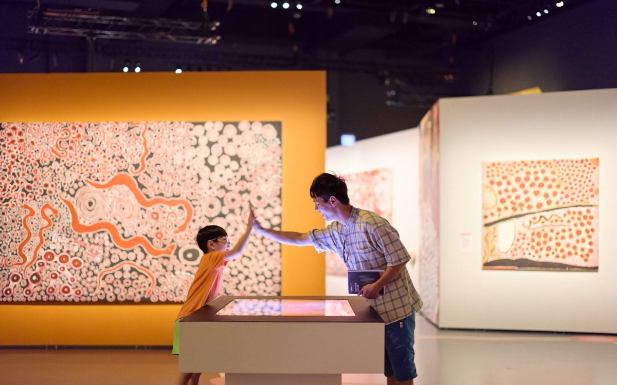 ‘Spinifex People: art and stories of Pila Nguru’ at WA Museum Boola Bardip. A kid high fiving an adult inside an exhibition with orange and white Aboriginal dot paintings.