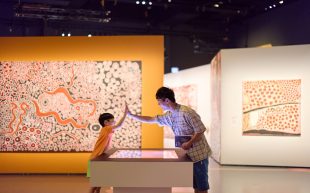 ‘Spinifex People: art and stories of Pila Nguru’ at WA Museum Boola Bardip. A kid high fiving an adult inside an exhibition with orange and white Aboriginal dot paintings.