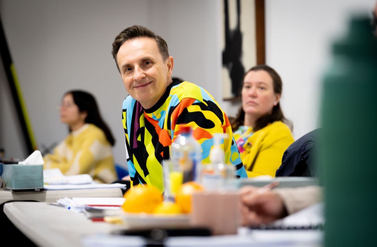 Mitchell Butel in the rehearsal room for STCSA's 2024 production 'The Questions'. Butel is a fair-skinned man aged 54, clean shaven and with neatly styled short brown hair. He wears a brightly-patterned jumper and is sitting at a table, leaning forward to smile for the camera.