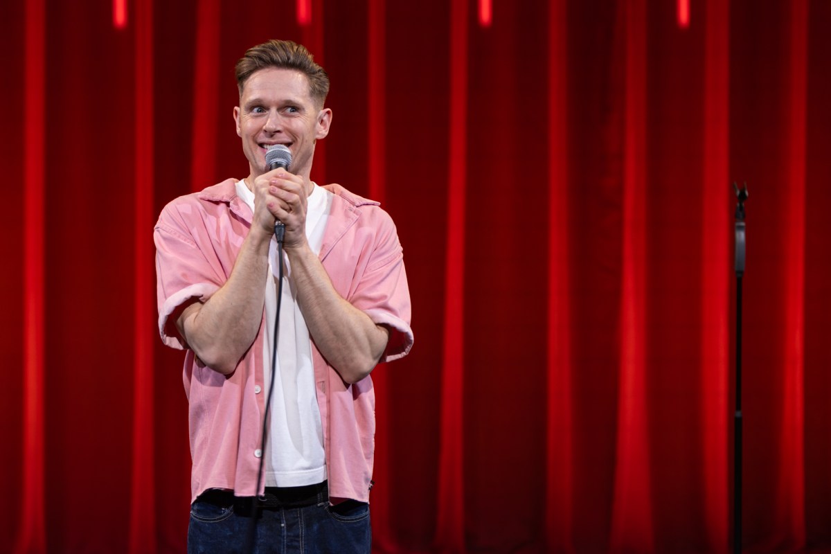 Samuel Barnett, a young man in a white t-shirt and pink short sleeve shirt is clutching a microphone on stage. The curtains behind him are red.