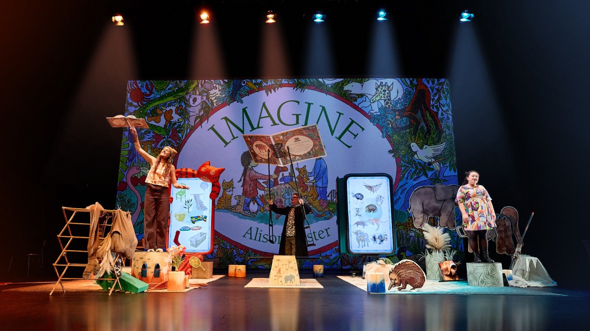 A large copy of Alison Lester's picture book 'Imagine Live' is placed in the middle of the stage, with two actors between it and various paraphrenalia from the book.