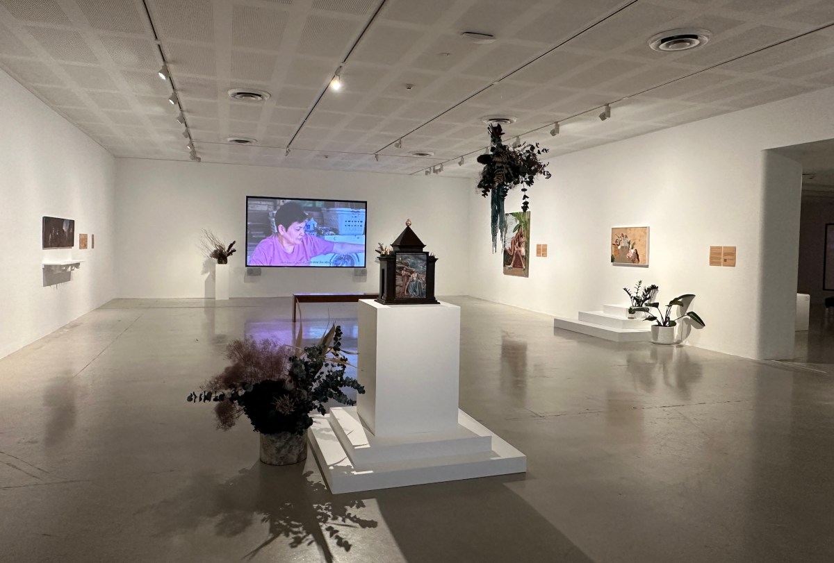 Gallery view of exhibition by Marikit Santiago. Inside a space with white walls and gray concrete floor are several pots of plants, paintings on walls, and a video on the far wall.