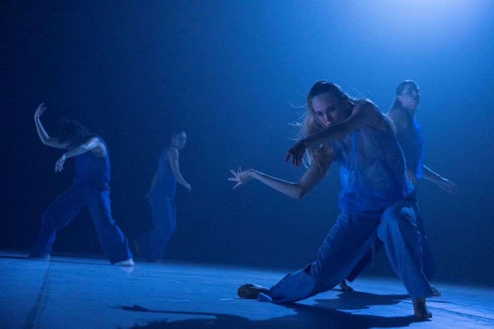 A sequence from Tra Mi Dinh’s 'Somewhere between ten and fourteen', which features in Sydney Dance Company's 2025 season. The photo depicts four barefoot dancers in blue coveralls dancing on a blue-lit stage. The dancer on the far left has their legs spread, their right arm raised and their left arm extended horizontally; the dancer on the right, who is the focus of the focus, stands in a half-crouch, her arms extended to the right and her right foot bent so that the side of the foot rather than her sole is touching the stage.