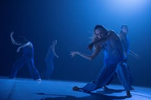 A sequence from Tra Mi Dinh’s 'Somewhere between ten and fourteen', which features in Sydney Dance Company's 2025 season. The photo depicts four barefoot dancers in blue coveralls dancing on a blue-lit stage. The dancer on the far left has their legs spread, their right arm raised and their left arm extended horizontally; the dancer on the right, who is the focus of the focus, stands in a half-crouch, her arms extended to the right and her right foot bent so that the side of the foot rather than her sole is touching the stage.