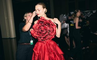 Model and crew backstage for the Melbourne Fashion Festival. A tall female model with pale skin and red lips is wearing a red satin dress with a large floral feature over her chest, while the crew adjusts something around her ear.