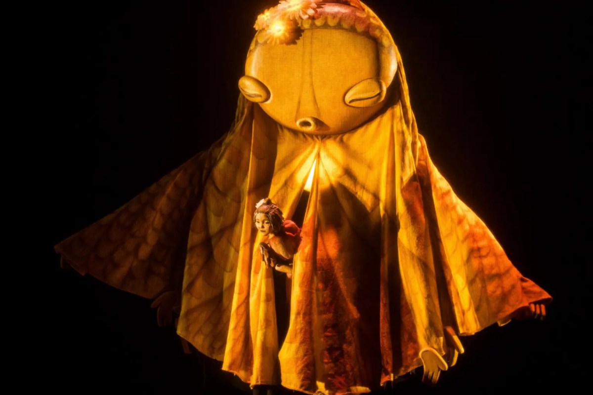 Spare Parts Puppet Theatre's 2025 season includes the world premiere of a new puppet musical, 'Hometown'. Pictured: a woman is peeping through the folds of a large, oversized puppet. The light on them both is golden; a scene from Spare Parts' 2025 production, 'Hometown.'