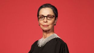 Australian actor Heather Mitchell as Ruth Bader Ginsburg in ‘RBG: Of Many, One’ at HOTA from 3-5 April 2025. A fair-skinned woman, dressed as member of the American judicary, stares somewhat aloofly at the camera. Behind her is a salmon-pink background.