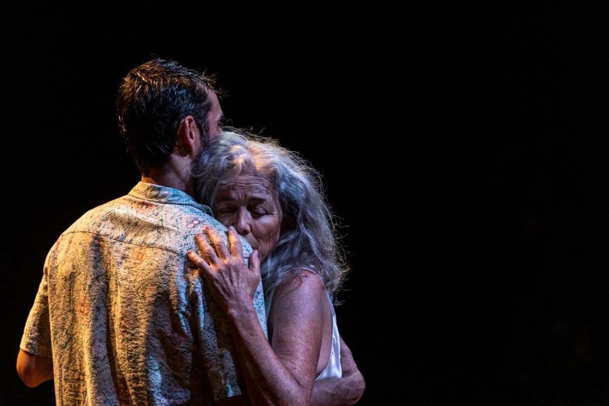 A man (Borja Maestre) and a woman (Sarah Peirse) are hugging. They are in spotlight. There is darkness around them.