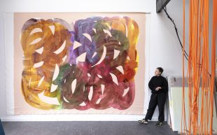 A figure standing next to a large unstretched canvas on a white wall, depicting thick circular strokes in yellow, orange, green, purple and red.