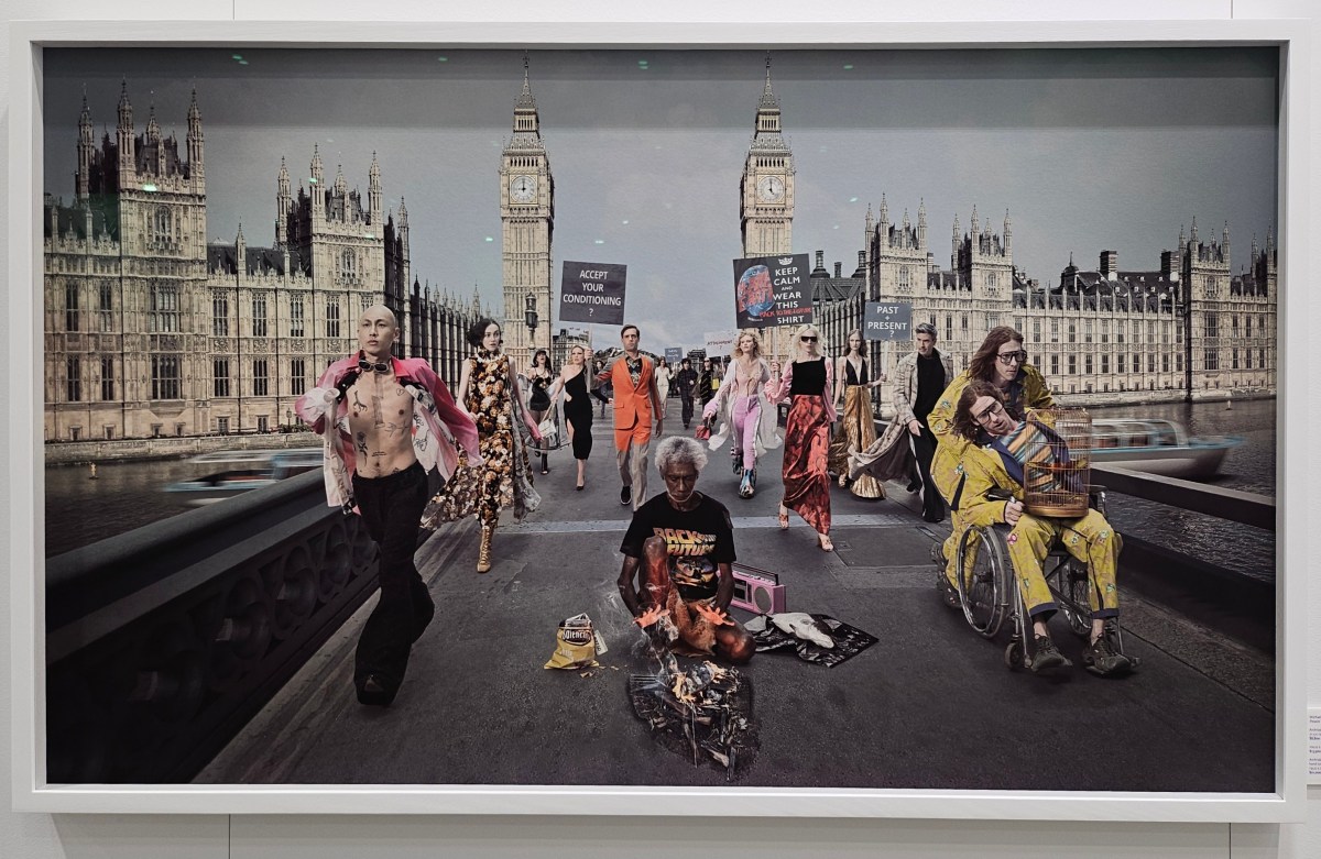 A photographic work showing a gray-haired Aboriginal dark wearing a ‘Back to the future’ tshirt sitting at a fire on the London bridge where people dressed stylishly are in a parade around him with signs saying “ACCEPT YOUR CONDITIONING?” And “PAST+PRESENT?’