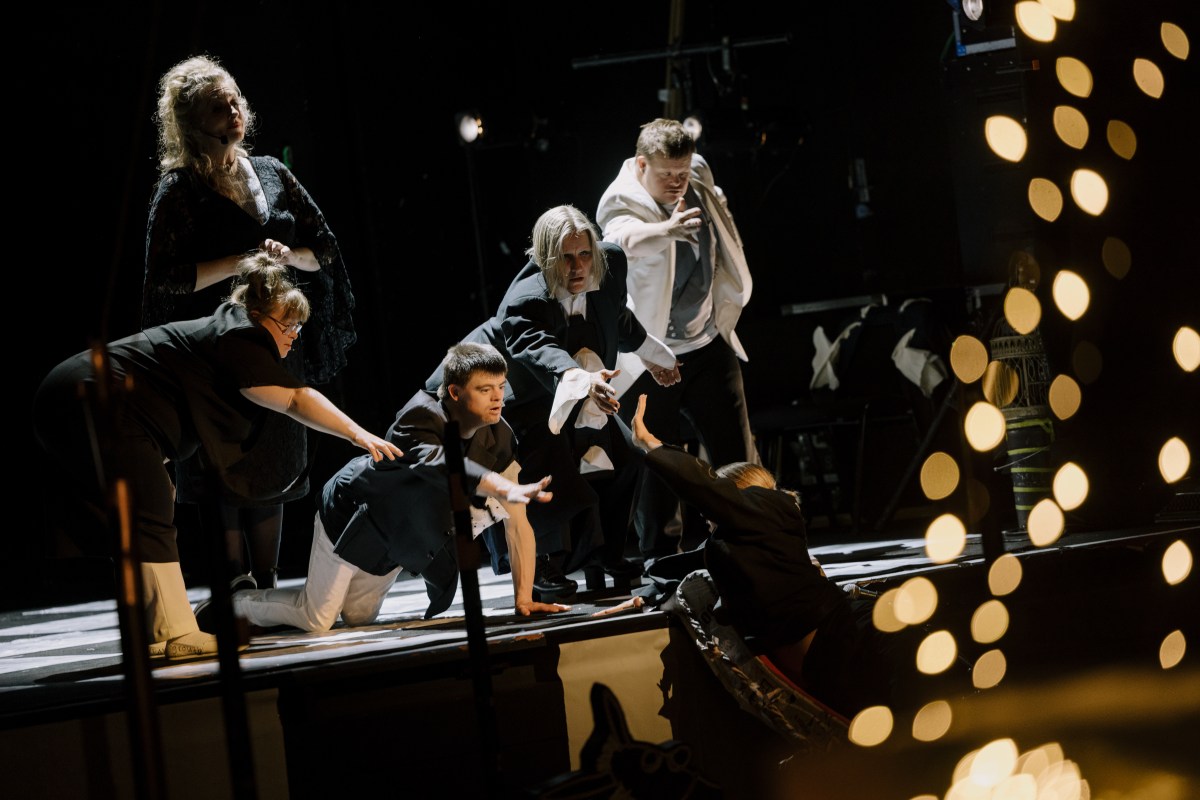 Three performers wearing black and white are reaching out to a third who is climbing on the stage.