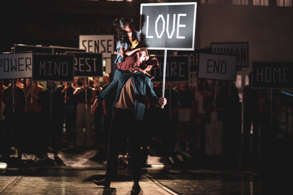 A man is standing, with a woman on top of him, He's holding up a sign with 'Love" on it. There is a crowd around them holding up signs with other words including 'Power', 'Mind', "rest' and "Sense'
